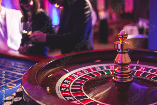 Vibrant casino table with roulette in motion, with casino chips, tokens, the hand of croupier, dollar bill money and a group of gambling rich wealthy people playing bet in the background