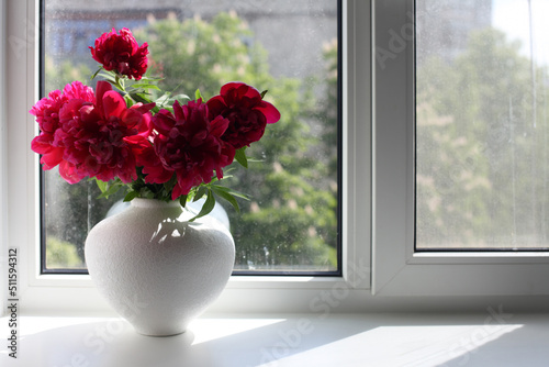 Red peony flower bouquet in white vase on window background. Minimalist still life.