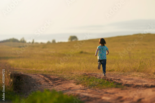 A small child runs along a dirt road in a meadow on the high bank of the river. There is a blurry image of a reservoir in the background.