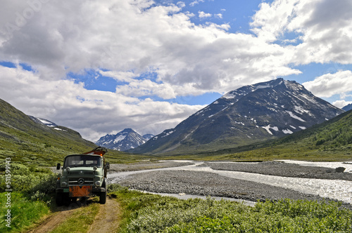 Obraz na płótnie High mountains with snow in summer, a valley with hills covered with grass and a stony river in Norway. An old truck stands on the river bank in Scandinavia. w salonie