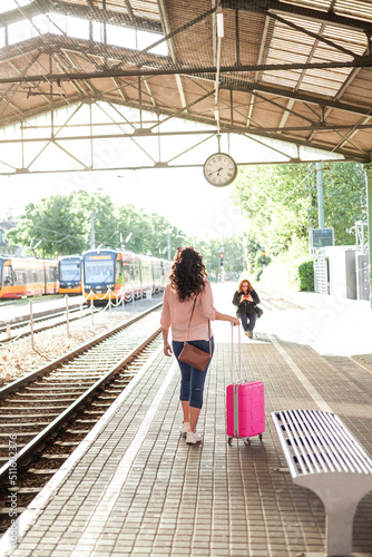 Young beautiful woman of 30-40 years old with a pink small suitcase is waiting for her train at the station. Curly model with casual clothes talking on the phone, drinking coffee to go. Travel.