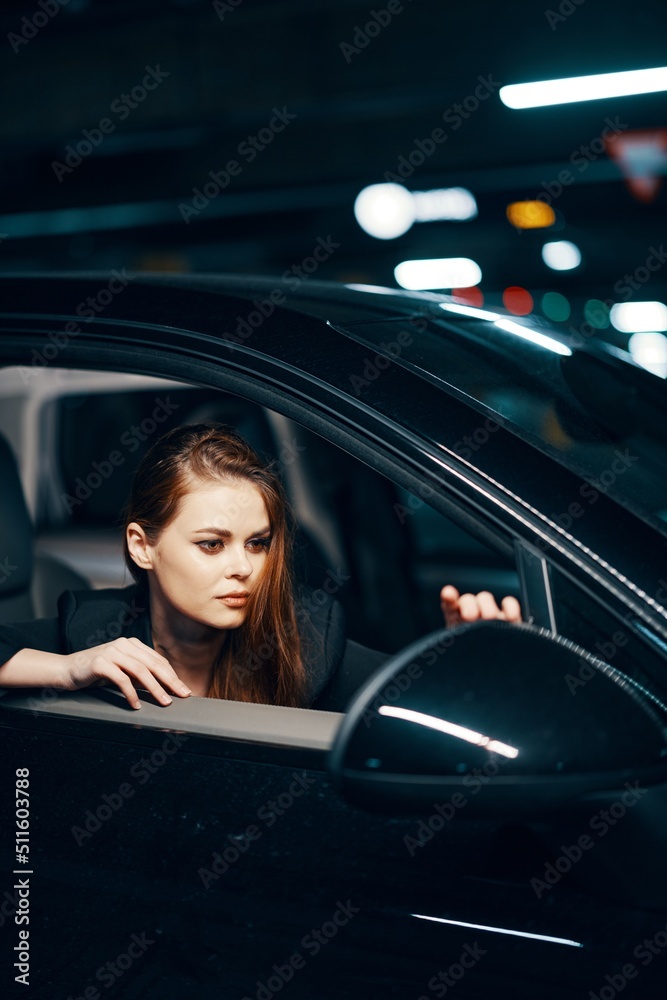 vertical photo from the side, at night, of a woman sitting in a black car and looking out of the window and reaching out to the side view mirror to correct it