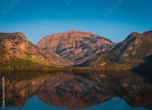 View of mountain range by the lake in Colorado, USA, at sunset; mountains reflect in calm water; blue sky in background