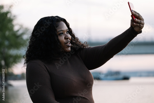 Pretty smiling dark skin interracial woman in elegant dress taking selfie, video call by smartphone, posing, gesturing by hand against seaside and sky. Multicultural person, diversity. Relax weekend