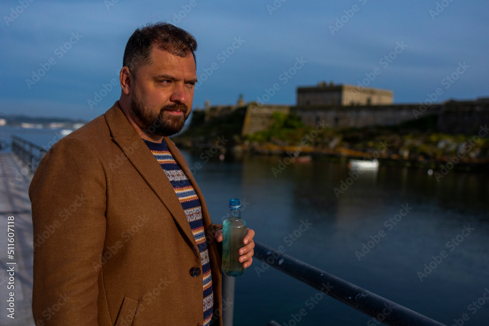 Fototapeta premium A man with a beard in a brown coat stands near the ocean and holds a bottle of water. The man is wearing a jacket with a pattern. A pensive look. In the background in the ocean is an ancient castle