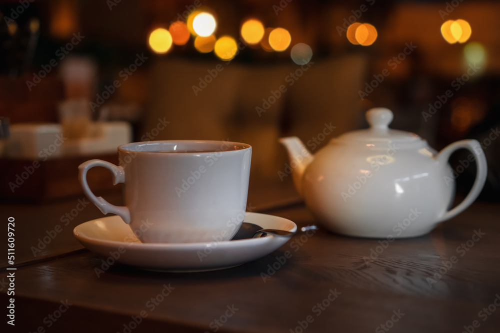 Fototapeta premium White cup with teapot. Pot standing on saucer in soft focus on naturally blurred background. Coffee, tea house, bokeh lights. The concept of a cozy pastime, tea ceremony