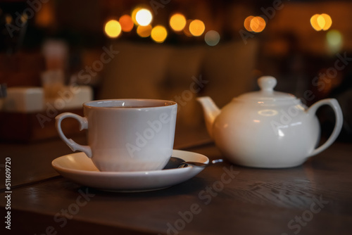 White cup with teapot. Pot standing on saucer in soft focus on naturally blurred background. Coffee, tea house, bokeh lights. The concept of a cozy pastime, tea ceremony