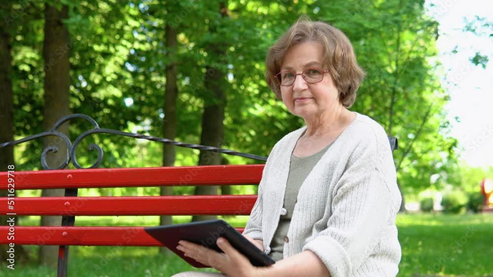 Elderly woman with a tablet in her hands sits on a bench, surfing the internet and researching news on social networks Elderly woman with a tablet in her hands sits on a bench, surfing the internet and researching news on social networks