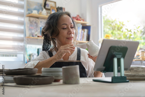 Small ceramic studio owner holding pottery earthenware product in front of digital tablet while live streaming on internet for selling. Free Download Small ceramic studio owner holding pottery earthenware product in front of digital tablet while live streaming on internet for selling. - GFXLoad
