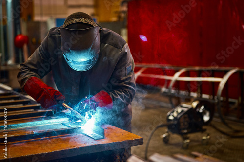Skilled worker in protective mask welds metal part at plant