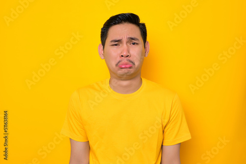 Unhappy young Asian man in casual t-shirt looking at camera with sad face isolated on yellow background. People lifestyle concept
