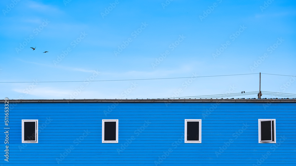 Minimal background of 4 Glass windows on blue wooden wall of row house with birds perching on electric cable lines against blue sky in widescreen view