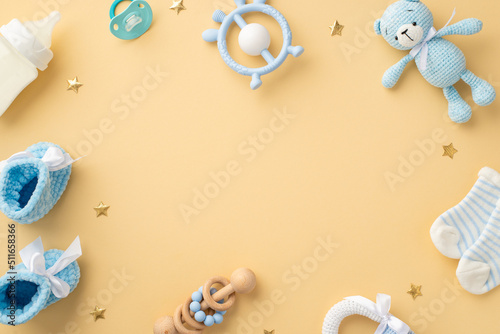 Baby concept. Top view photo of blue teddy-bear toy rattle socks milk bottle teether soother knitted shoes and gold stars on isolated pastel beige background with empty space in the middle photo