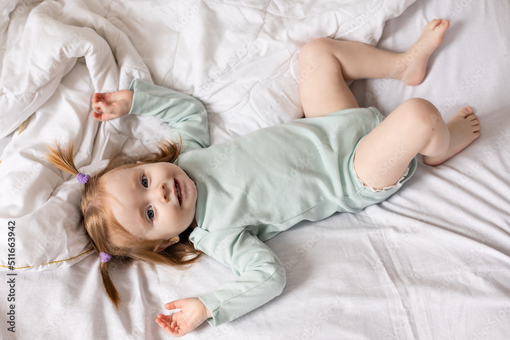 little girl in a light green bodysuit is lying in bed made with white bed linen