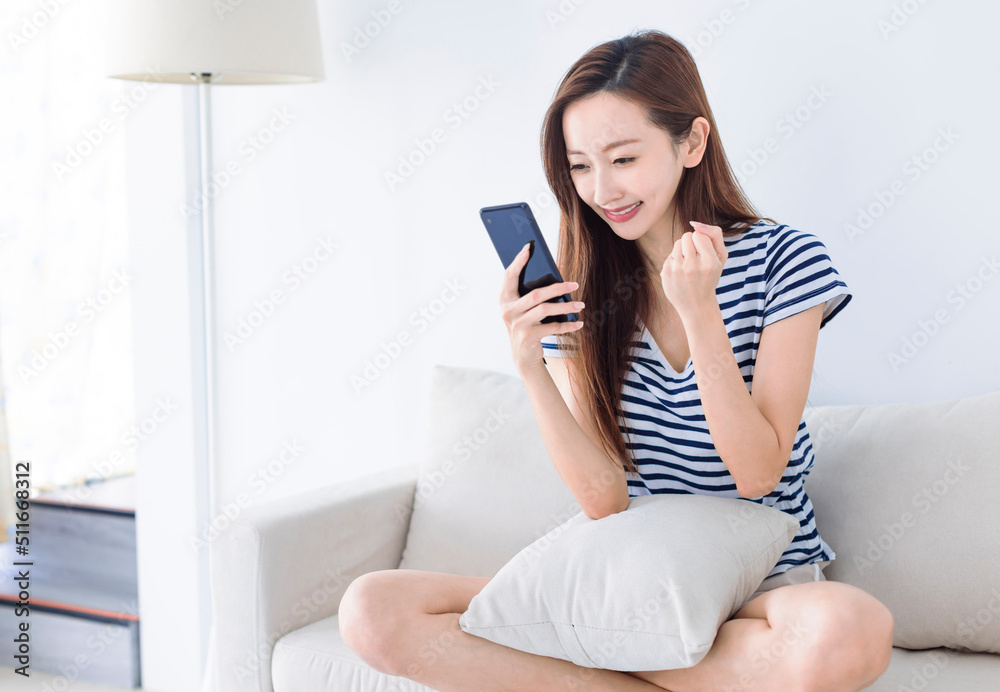 excited young woman looking at her smart phone and smiling while sitting on the sofa at home excited young woman looking at her smart phone and smiling while sitting on the sofa at home