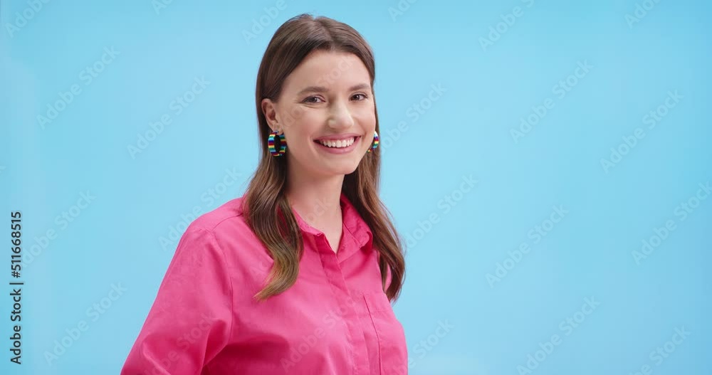 Portrait of young happy beautiful female looking at camera and smiling in positive mood posing on blue background in studio. Smiling Caucasian pretty woman in pink blouse with smile on face, close up Portrait of young happy beautiful female looking at camera and smiling in positive mood posing on blue background in studio. Smiling Caucasian pretty woman in pink blouse with smile on face, close up