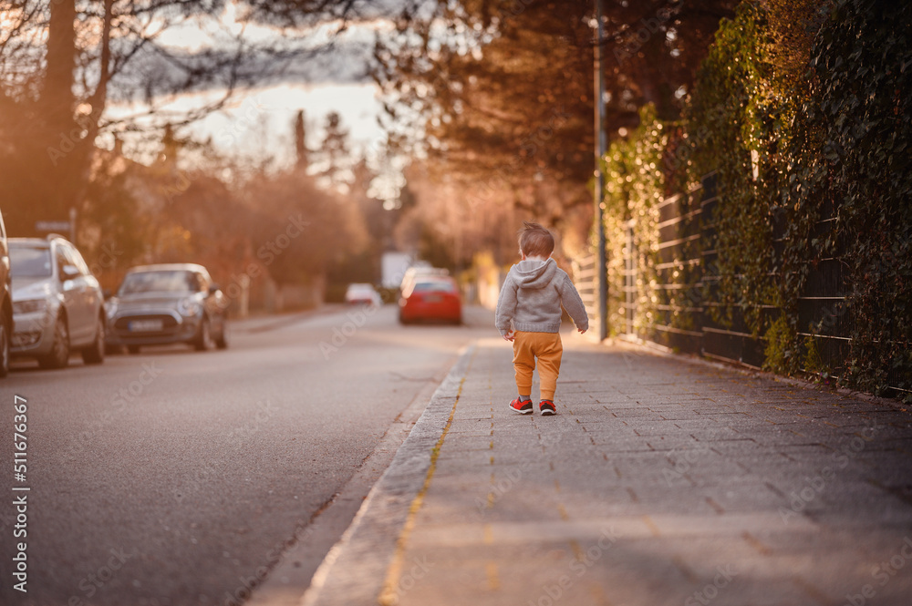 Little toddler boy in spring outwear walking alone on street in evening sunlight