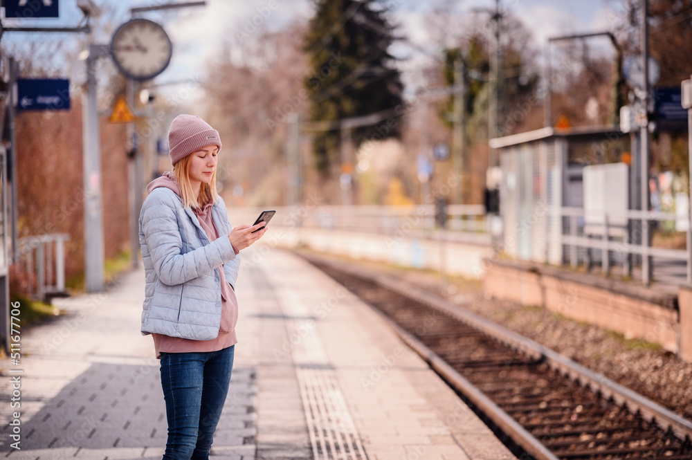 Young blonde woman in hat standing alone on railway station speaking by smartphone, waiting train