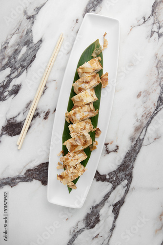 set of bonito rolls with grilled salmon, avocado, tuna shavings and green bamboo leaf in a white ceramic plate with chopstick on a bright textured marble background, top view