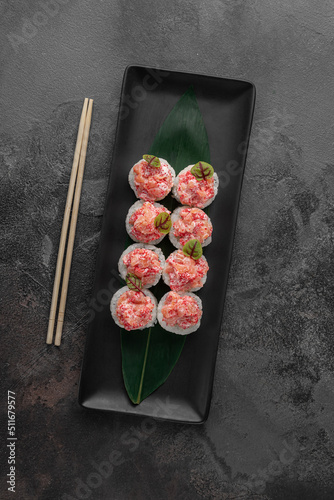 set of rolls with salmon, red flying fish caviar and green bamboo leaf in a black ceramic plate with chopstick on a dark gray textured background, top view