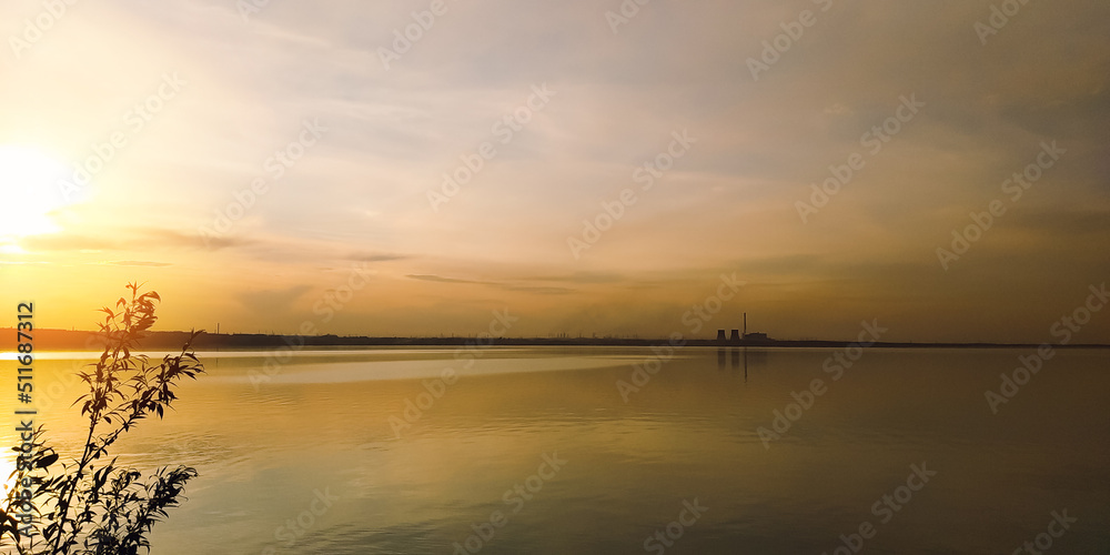 The sun at sunset over a branch of a large lake and factories on the horizon