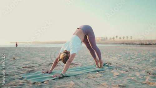Beautiful shot of stylish woman exercising yoga on fitness mat by the sea. Blond yogi girl dressed in sportswear doing downward dog pose on the beach
