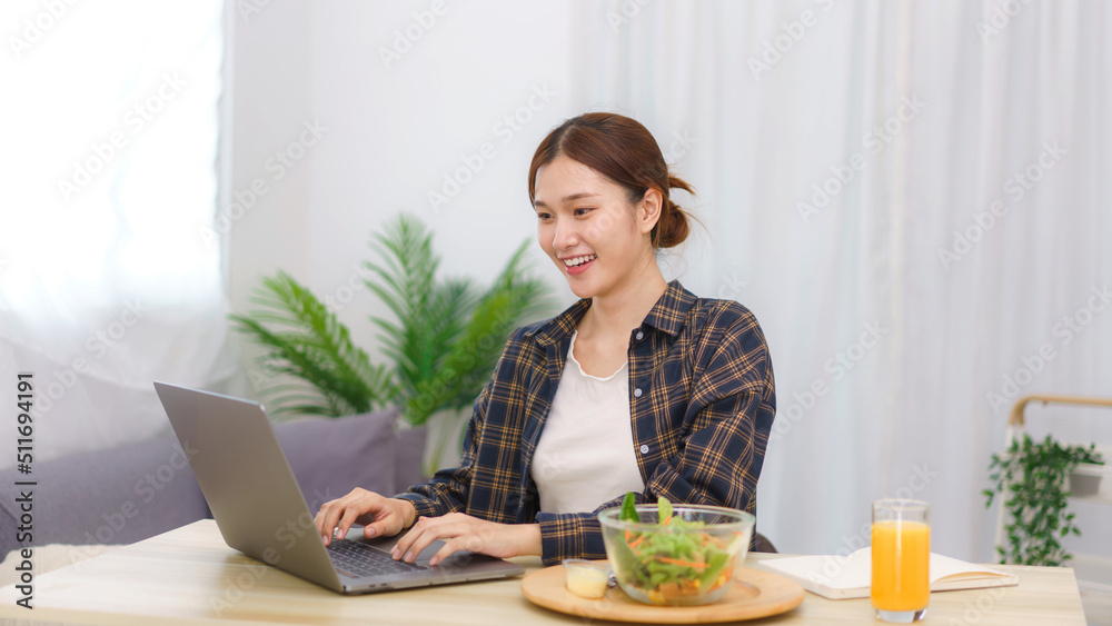 Lifestyle in living room concept, Young Asian woman eating vegetable salad and working on laptop