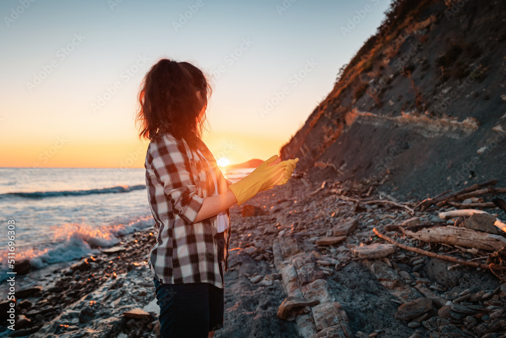 Woman in protective gloves looks to the sunset. Copy space. Wild beach and ocean in the background. Back view. The concept of Earth Day
