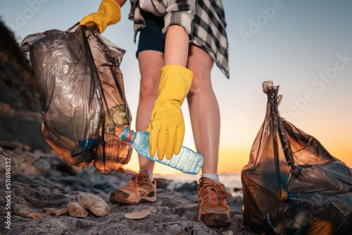 Female volunteer holding two polyethylene bags and picks up a plastic bottle on the beach. Close up of hand. Low angle view. The concept of cleaning the coastal zone
