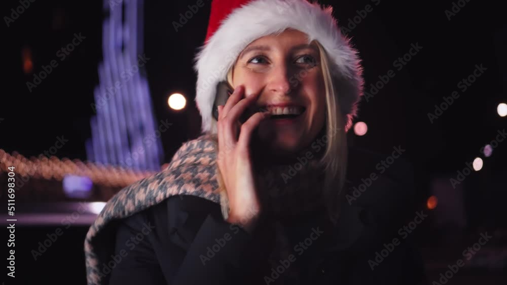 close-up of a white woman chatting on her smartphone. A pretty woman in a Santa hat. a girl is talking on the phone and smiling against the background of city lights on Christmas or New Year's Day. close-up of a white woman chatting on her smartphone. A pretty woman in a Santa hat. a girl is talking on the phone and smiling against the background of city lights on Christmas or New Year's Day.