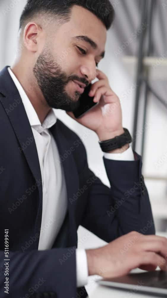 Office routine. Multitask man. Mobile communication. Work inspiration. Handsome business guy in elegant suit talking smartphone typing laptop sitting light room interior vertical. Office routine. Multitask man. Mobile communication. Work inspiration. Handsome business guy in elegant suit talking smartphone typing laptop sitting light room interior vertical.