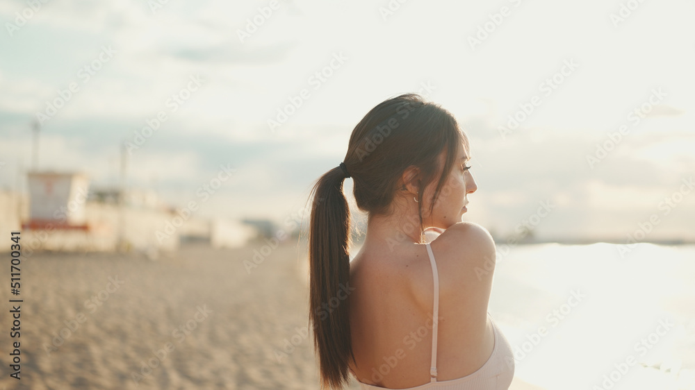 oung athletic woman with long ponytail wearing beige sports top is stretching on the beach on modern city background. - obrazy, fototapety, plakaty 