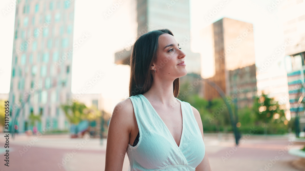 Lovely young woman with freckles and dark loose hair wearing white top and jeans is enjoying the view of modern city. Beautiful girl with long eyelashes looks at the upper floors of building