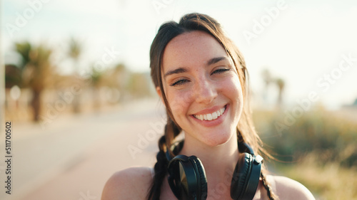 Fotografie Cute young athletic woman with braided pigtail wearing beige sports top with headphones stands on the embankment