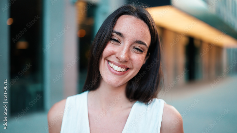 Close-up of young woman with freckles and dark loose hair and long eyelashes wearing white top looking straight at the camera. Beautiful girl on modern city background