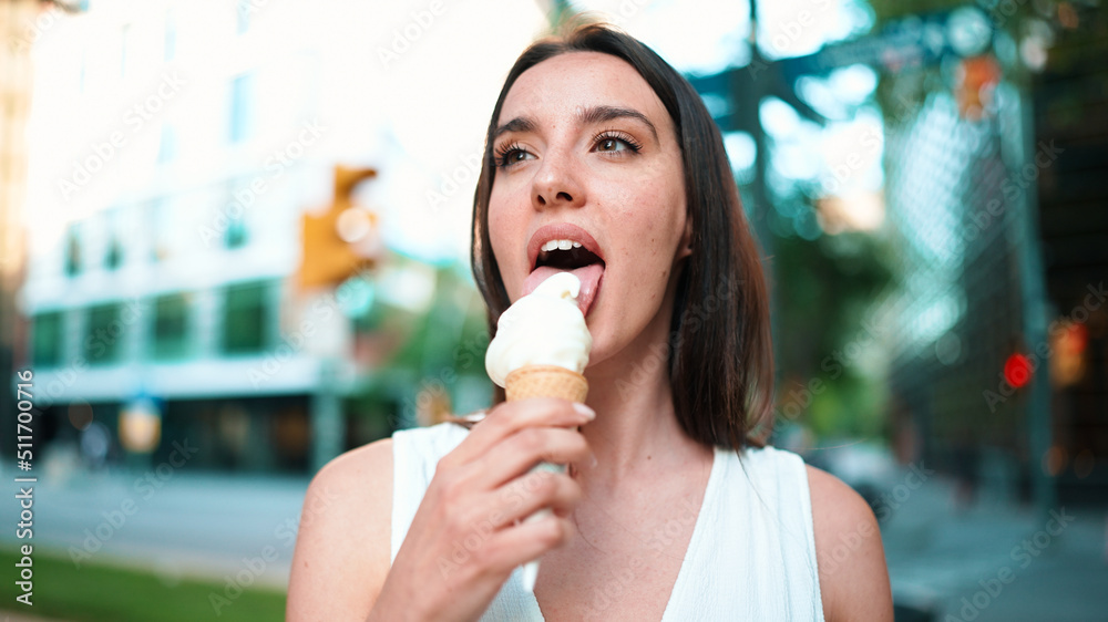 Close-up beautiful woman with freckles and dark loose hair wearing white top testing ice cream. Cute girl enjoys ice cream on modern city background