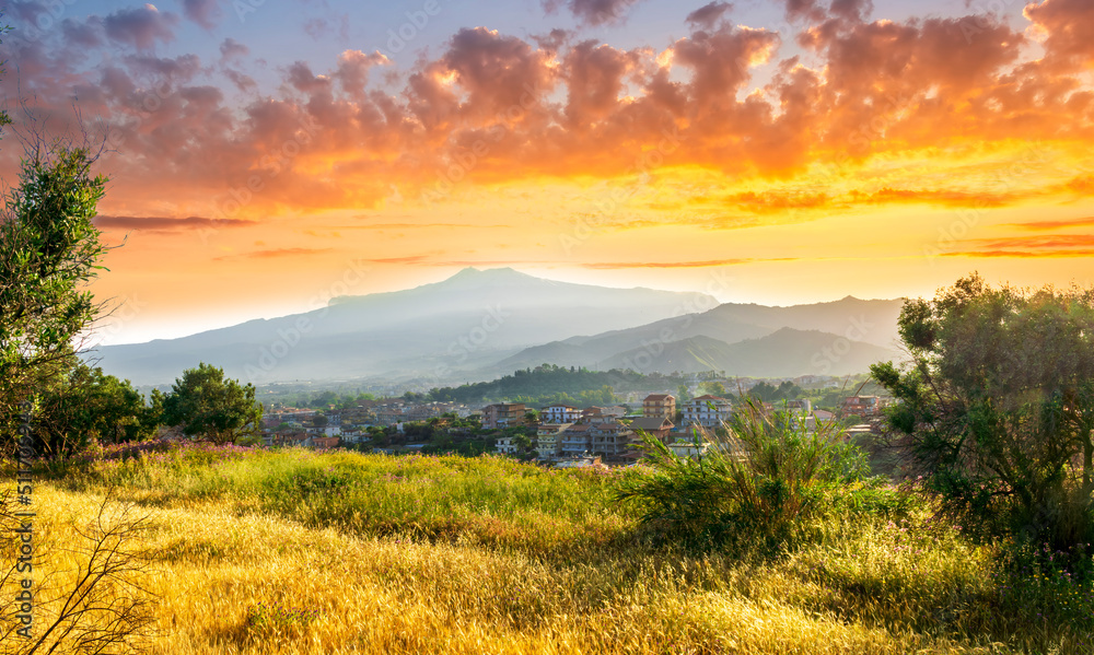 view from hill with golden grass and green bushes to a valley town with majectic mountains and scenic cloudy sunset on background
