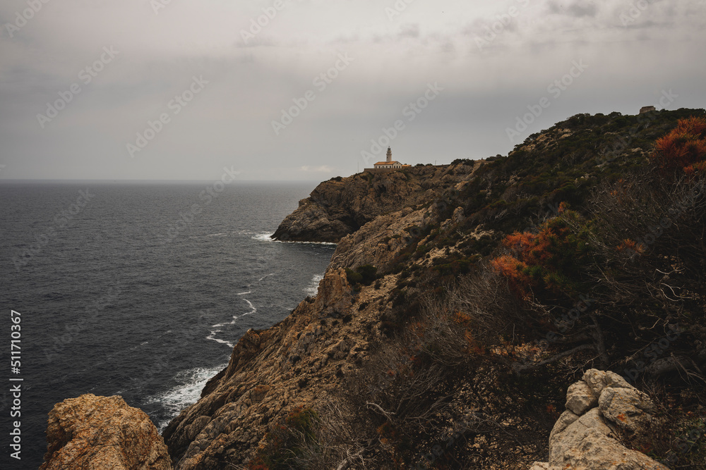 Biała latarnia morska na skalistym klifie w pochmurny dzień. Latarnia położona jest na wschodnim wybrzeżu Majorki. 