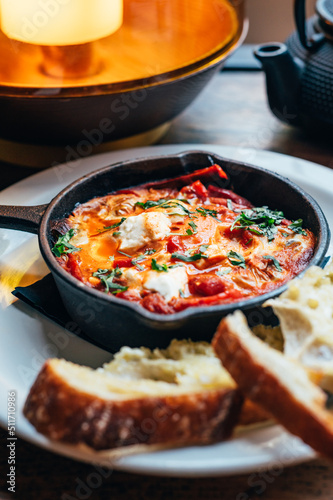 Freshly made shakshuka with spiced tomato  red pepper  feta  egg  coriander and pieces of homemade sourdough  served in iron pan  healthy vegetarian breakfast
