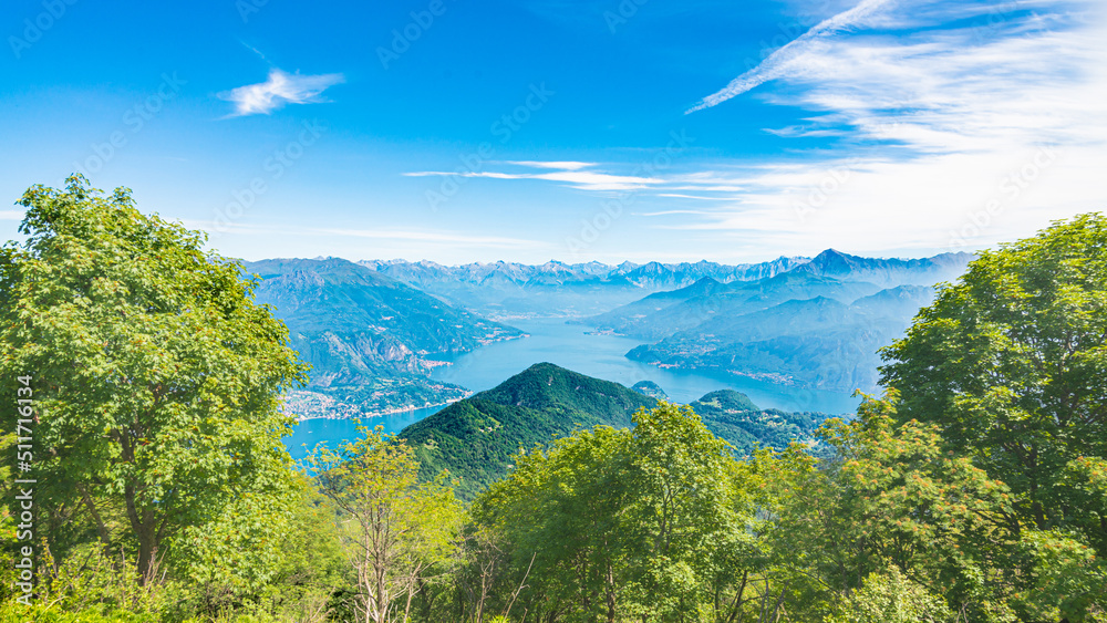 Fototapeta premium Aerial view of Lake Como, Lombardy, Italy, from a viewpoint on the italian alps. Green trees in the foreground. Blue sky with white clouds on the background.