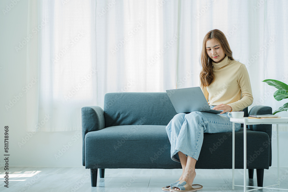 A freelance Asian woman working on a laptop checks social media while sitting on the sofa relaxing in her living room at home. concept of lifestyle for women