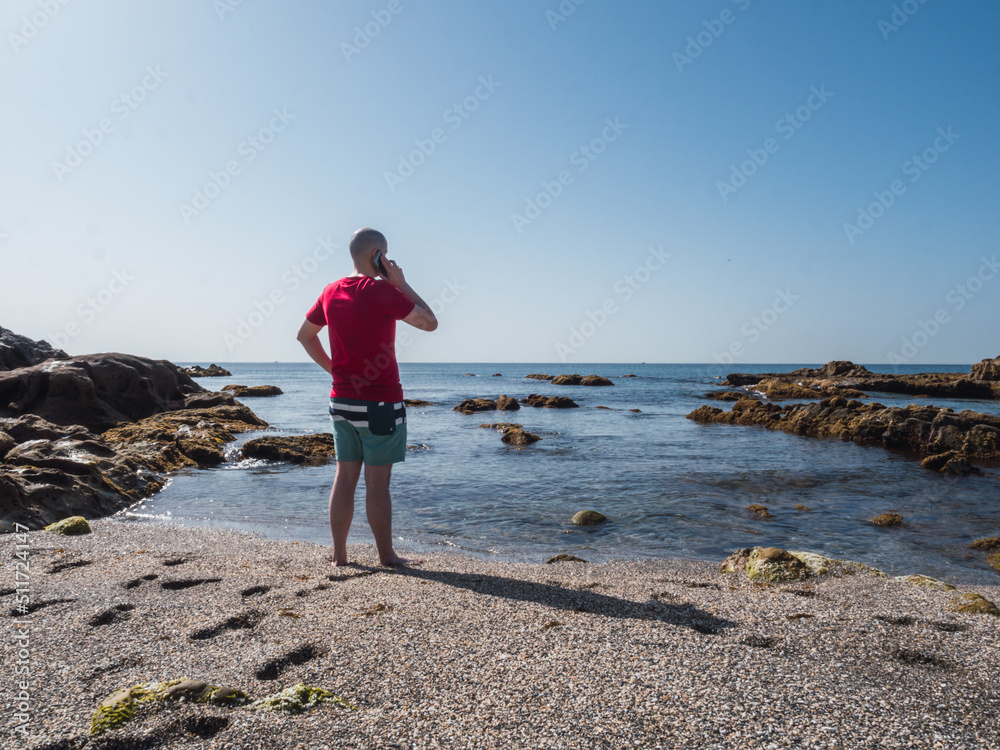 Caucasian tourist with red t-shirt recently arrived at the beach calling family from Mediterranean beach with clear sky and copy space