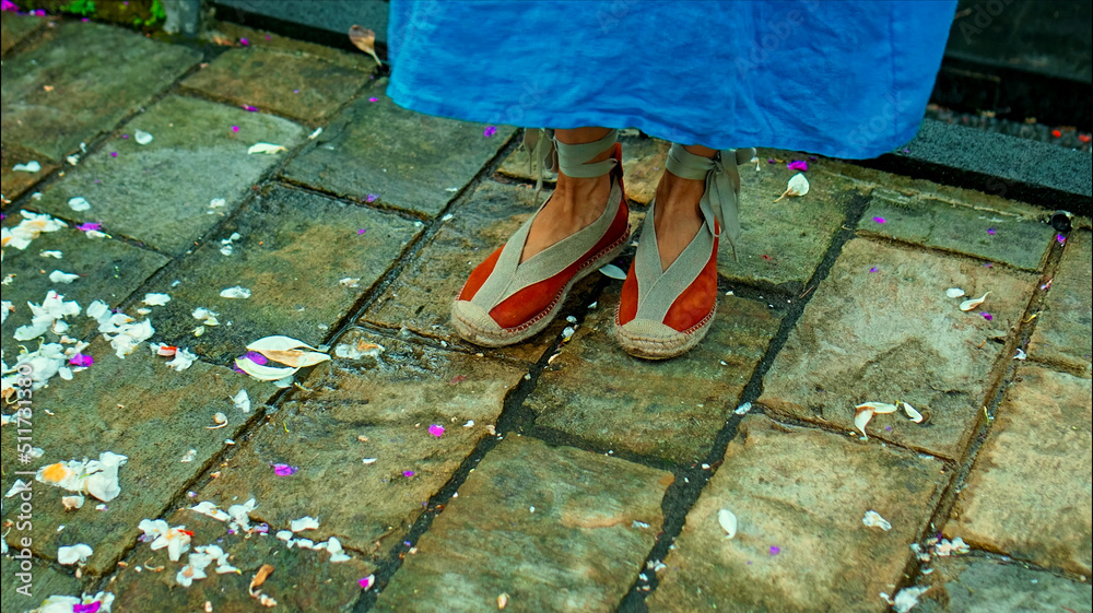 Legs of a European girl in a blue dress walking in shoes along the road with antique stonework along a green park with flowers