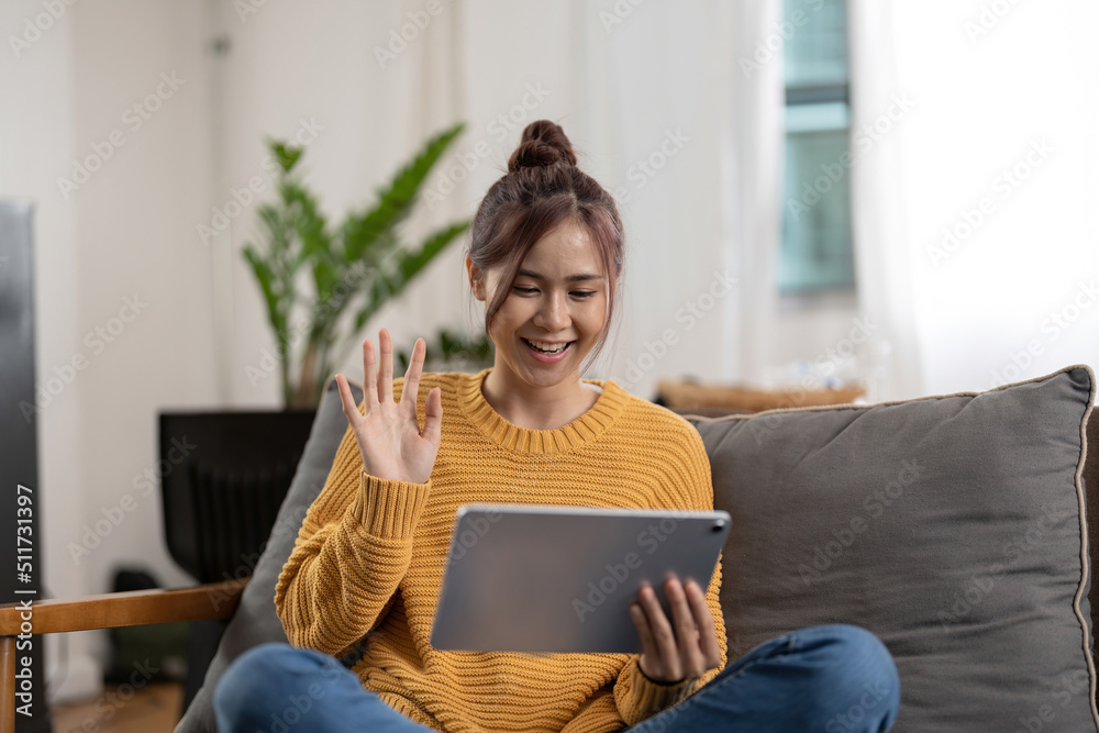 Happy smiling asian young woman in yellow sweater with tablet pc computer having video call and waving hand at home Happy smiling asian young woman in yellow sweater with tablet pc computer having video call and waving hand at home