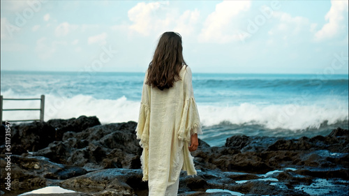 A young European girl with long black hair and a white robe walks to Water blow with cliffs and strong waves of seawater with clouds and blue sea