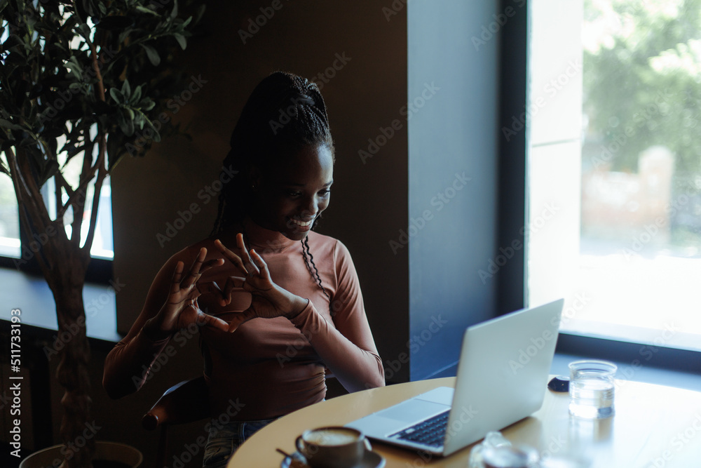 Happy smiling afro american woman showing heart with hands and talking on laptop online in coffee house. Black girl working remotely, freelance. Internet communication, video calling, love expression. Happy smiling afro american woman showing heart with hands and talking on laptop online in coffee house. Black girl working remotely, freelance. Internet communication, video calling, love expression.