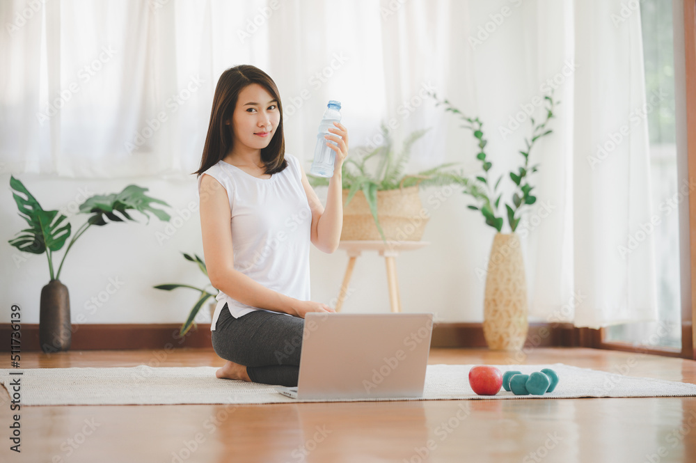 Healthy young Asian woman holding drinking water bottle while using laptop practicing yoga at home Healthy young Asian woman holding drinking water bottle while using laptop practicing yoga at home
