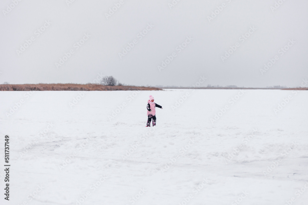 Child on background of winter landscape. Little girl in arm suit and hat with pompom stands on snow-covered ice in middle of lake.