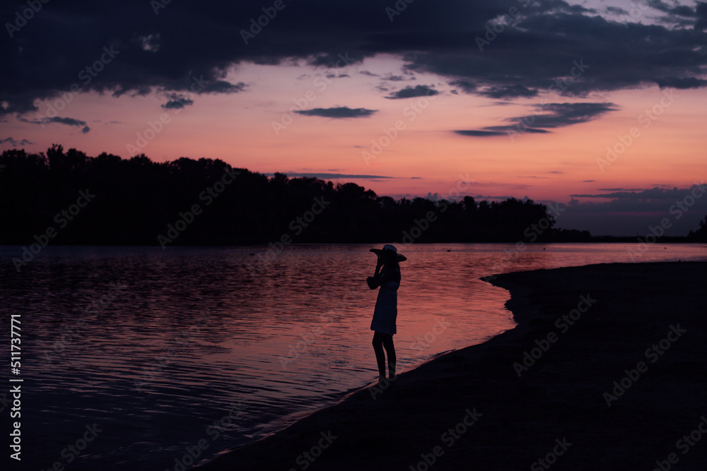 The silhouette of a woman in profile against the background of water and sunset. A slender young woman stands in a dress and hat on the beach and looks at the clouds and the picturesque landscape. 