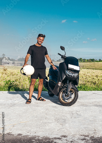 young man standing next to new black motorbike scooter around rice field in bali indonesia